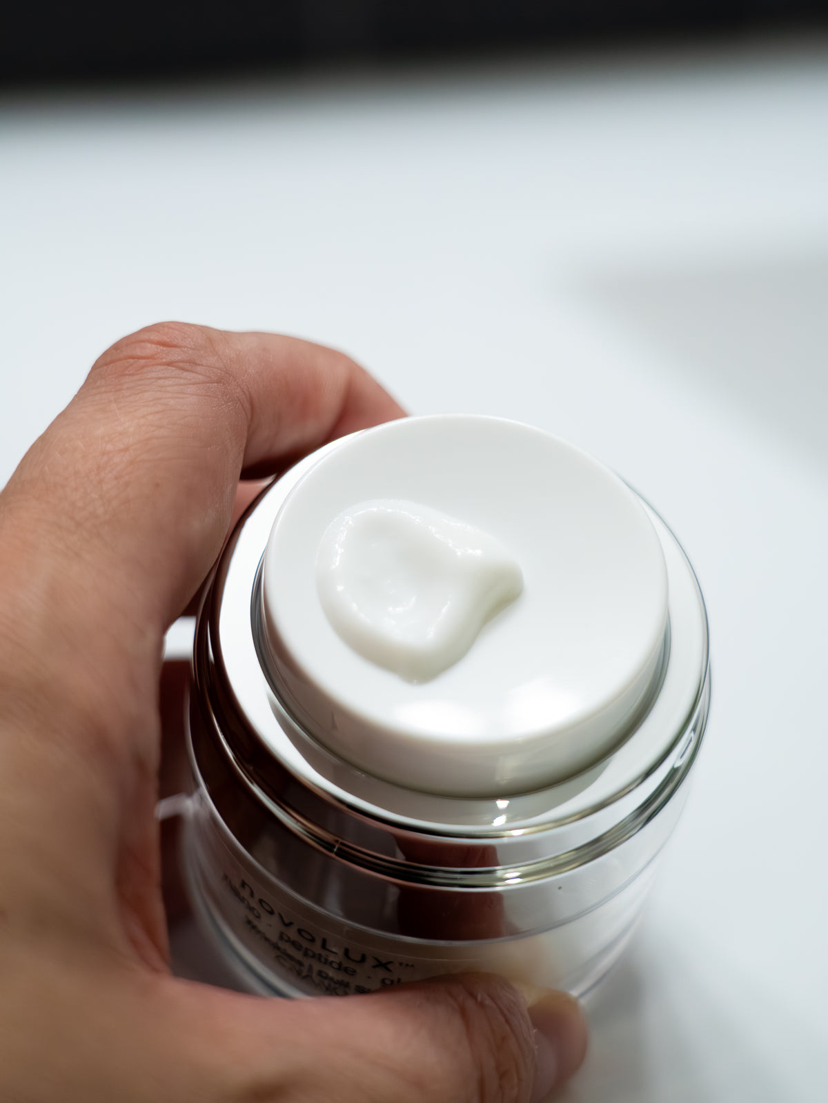 A hand holding a clear novoLUX jar with a white lid, showing a dollop of white cream on top. 
